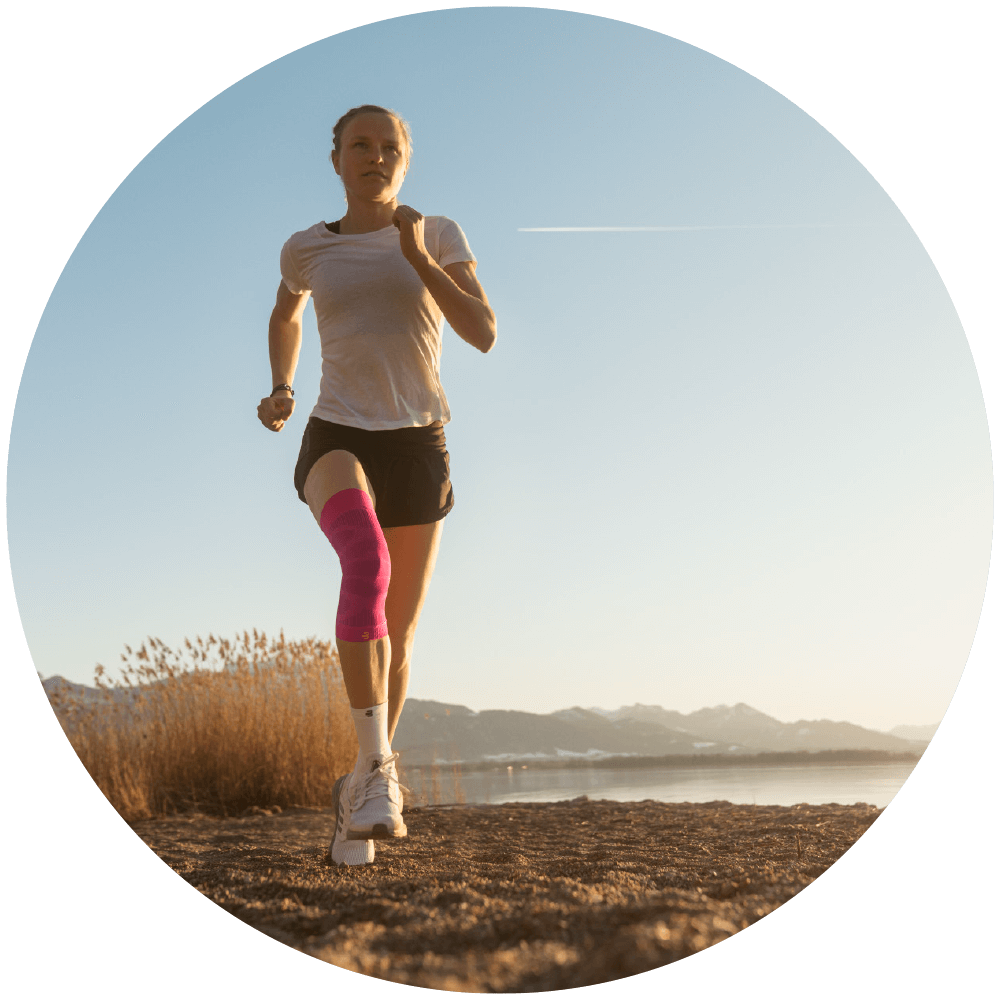 Improves Confidence Woman wearing Bauerfeind's Sports Compression Knee Support while running on a grassy trail near a lake.