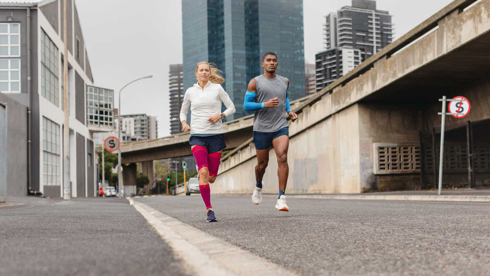 Two runners on an inner-city street while wearing the Sports Compression Sleeves. Two young adults running on an inner-city street while wearing Bauerfeind's Sports Compression Sleeves for the arm, upper leg and lower leg.