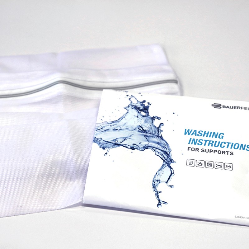 A Bauerfeind washing instructions booklet with a dynamic blue water splash graphic, placed beside a white fabric support on a light grey background.