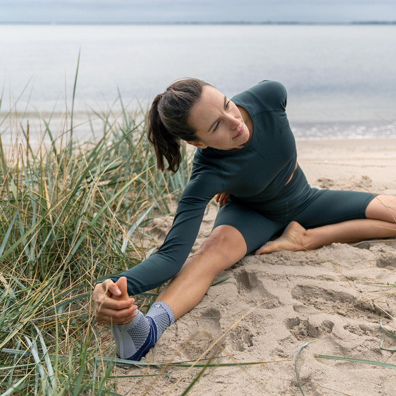 Yoga by the Lake: Wellness and Fitness Outdoors Woman practicing yoga on a sandy beach overlooking a serene lake, wearing a green workout outfit and a supportive ankle brace.