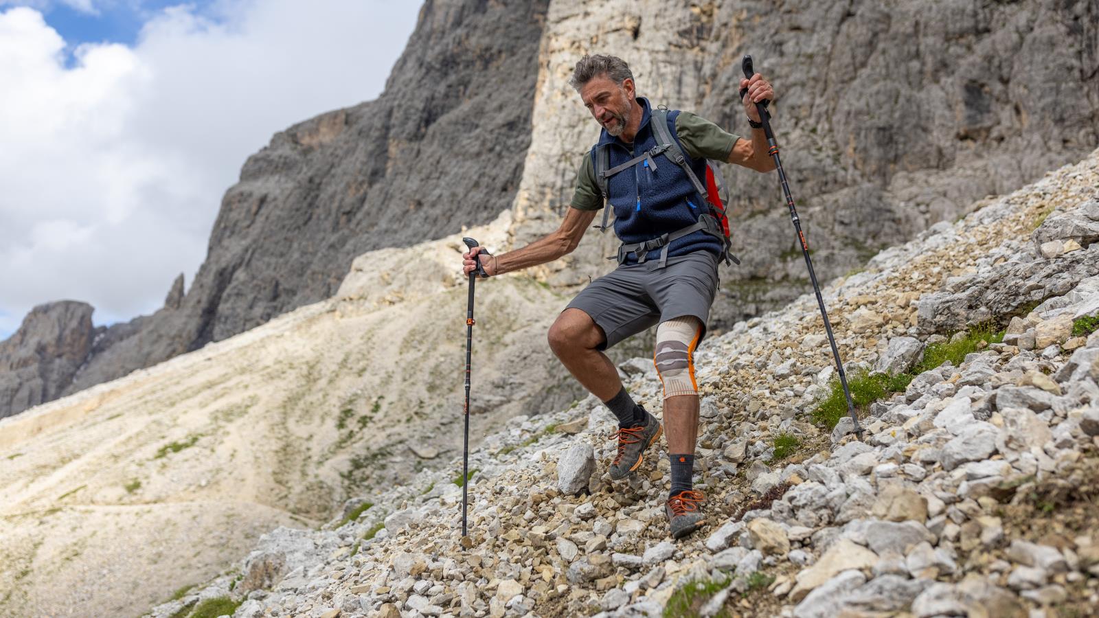 Man hiking downhill while wearing the Outdoor Knee Support. Man hiking downhill with trekking poles and hiking equipment while wearing Bauerfeind's Outdoor Knee Support.