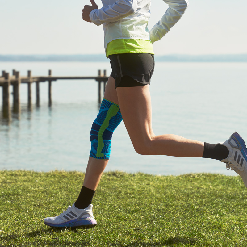 Woman wearing Bauerfeind's Sports Knee Support while running next to a lake.
