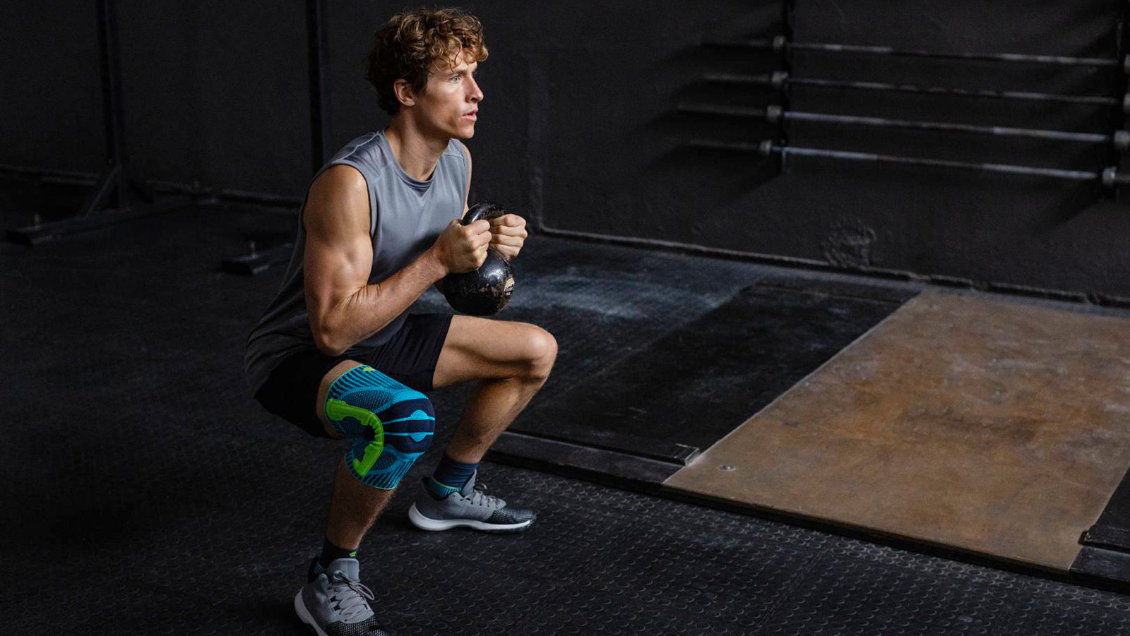 Man wearing the Sports Knee Support while performing kettlebell squats. Man wearing Bauerfeind's Sports Knee Support inside of a fitness center while performing kettlebell squats.