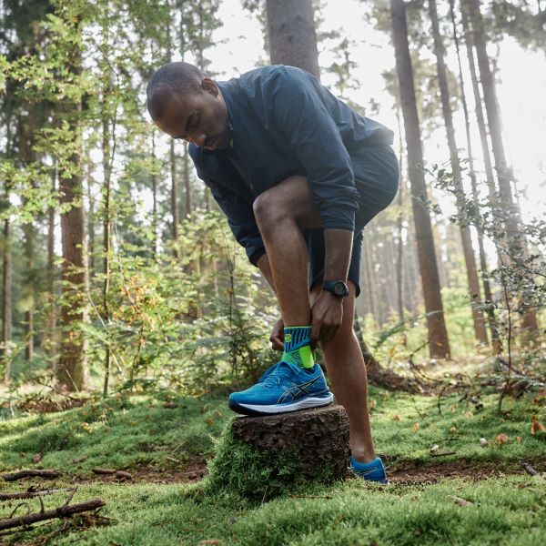 Man adjusting Bauerfeind's Sports Ankle Support before continuing to hike on a small dirt trail through the forest.
