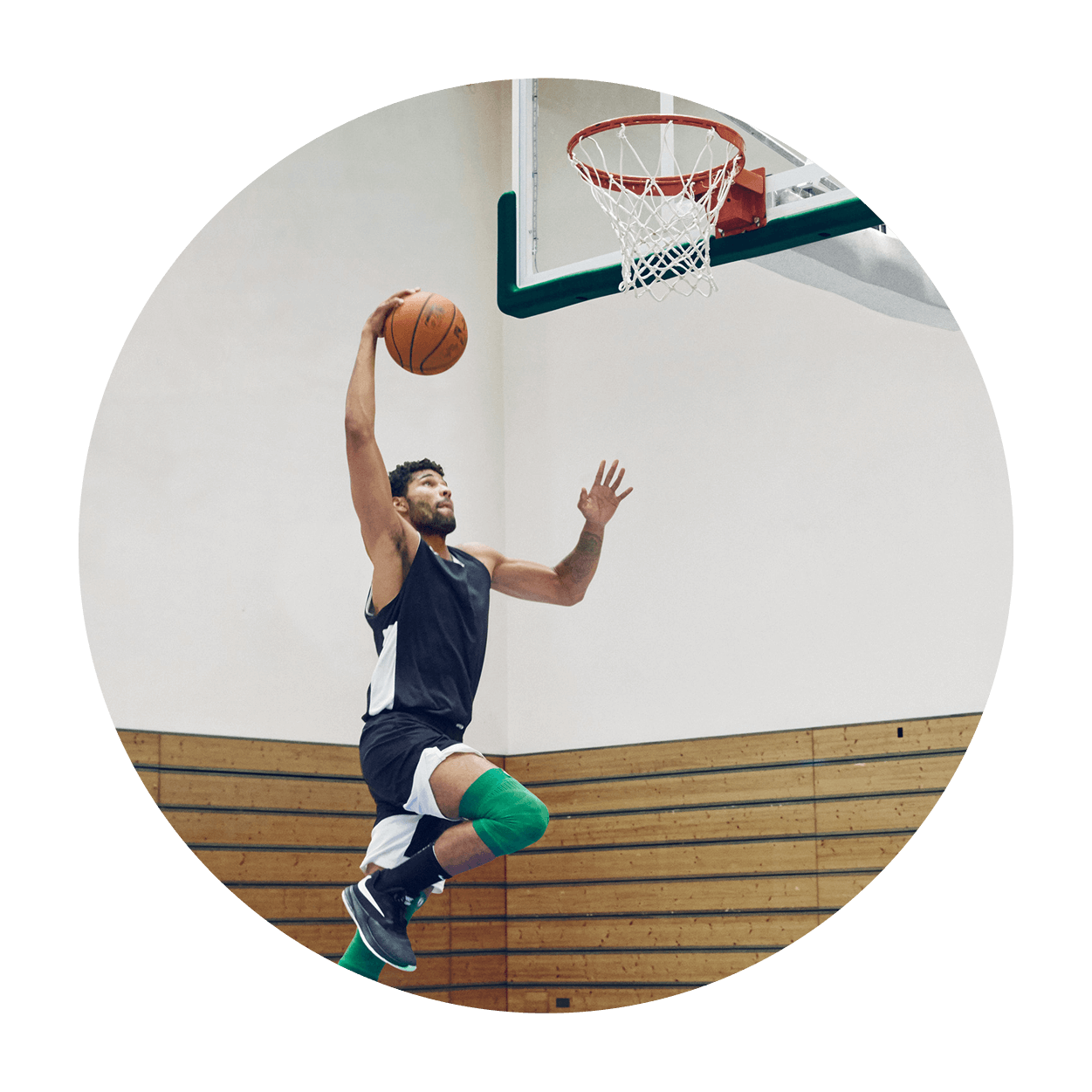 Athlete attempting a dunk while wearing the Sports Compression Knee Support NBA Celtics Edition. Athlete attempting a dunk on an indoor basketball court while wearing Bauerfeind's Sports Compression Knee Support NBA Celtics Edition.