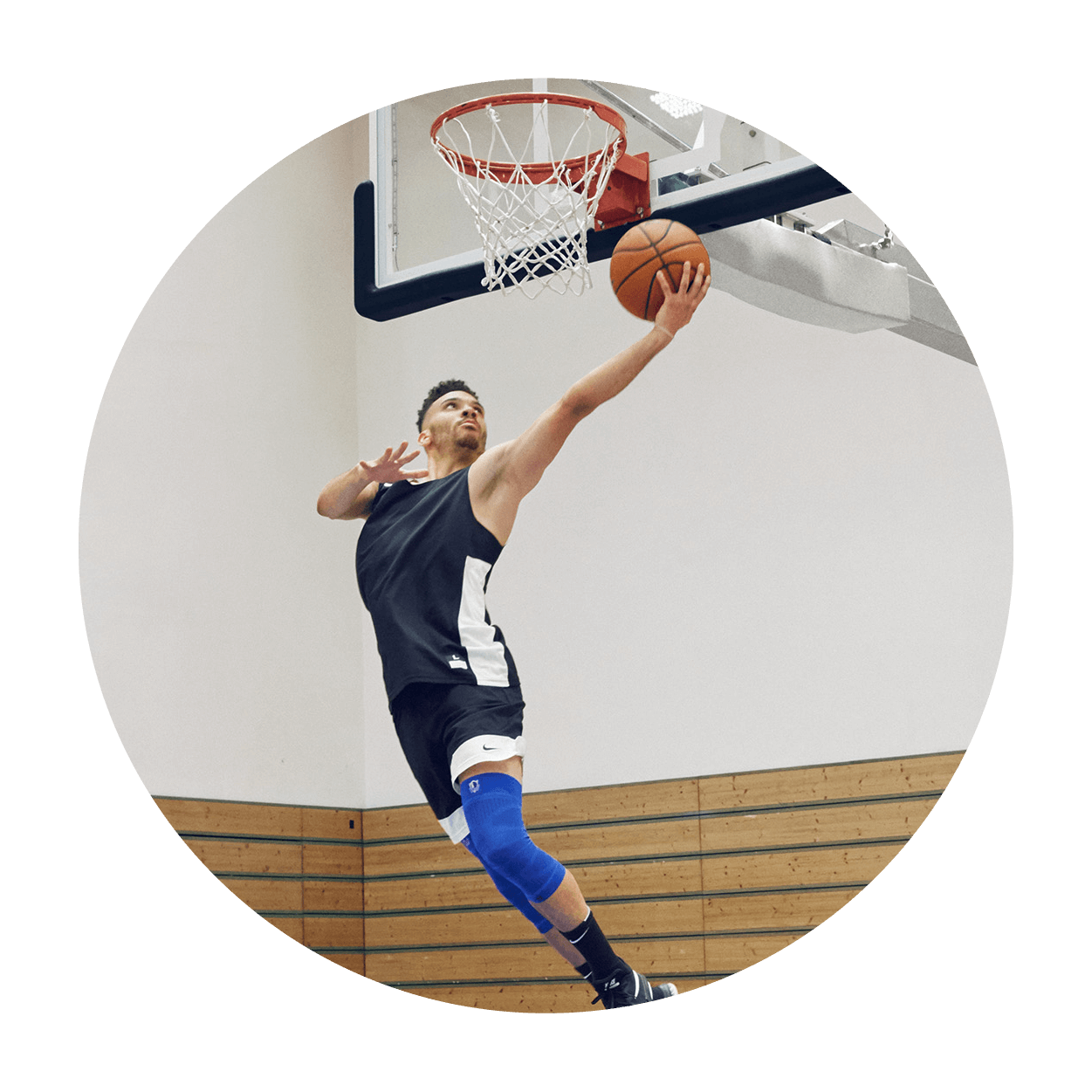 Athlete attempting a layup while wearing the Sports Compression Knee Support NBA Mavericks Edition. Athlete attempting a layup on an indoor basketball court while wearing Bauerfeind's Sports Compression Knee Support NBA Mavericks Edition.