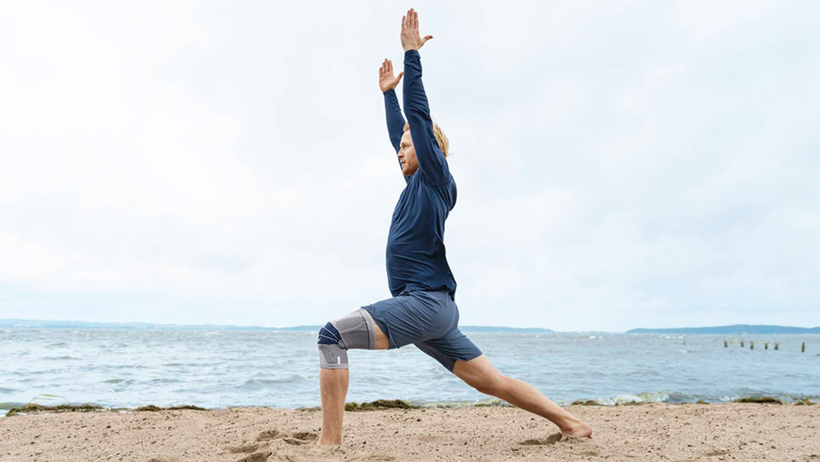 Man practicing yoga while wearing the GenuTrain on the beach. Man practicing yoga while wearing Bauerfeind's GenuTrain knee brace on the beach.
