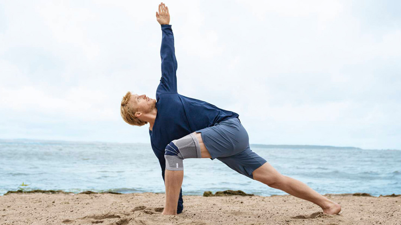 Man practicing yoga while wearing the GenuTrain on the beach. Man practicing yoga while wearing Bauerfeind's GenuTrain knee brace on the beach.