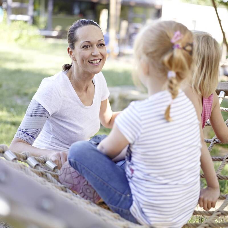 Woman wearing Bauerfeind's OmoTrain shoulder brace while playing at the park with two children.