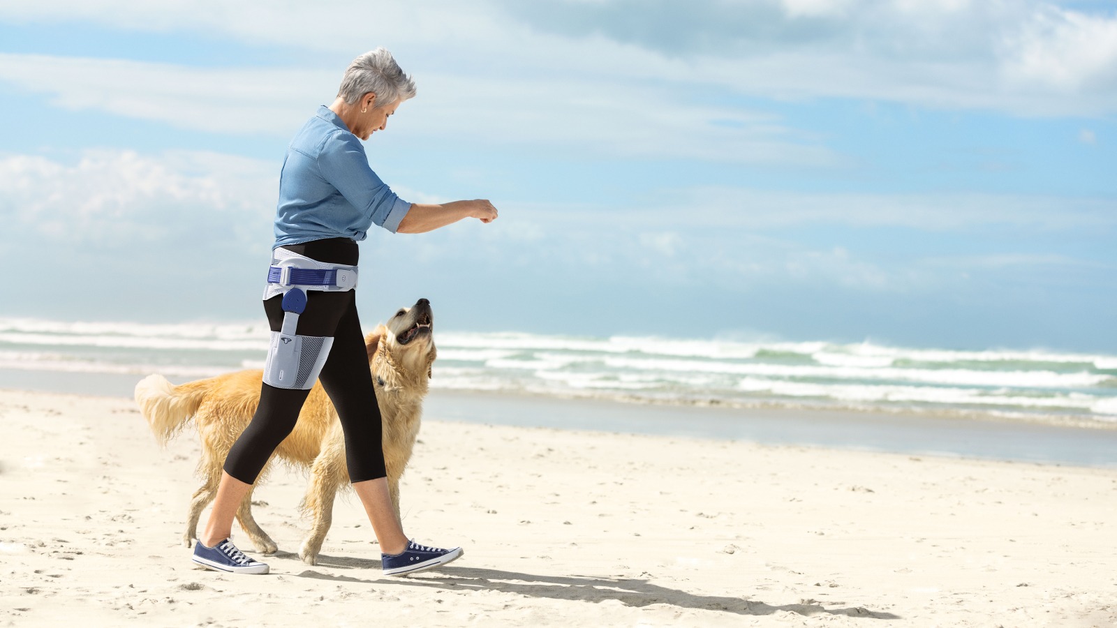 Woman wears Bauerfeind's CoxaTrain osteoarthritis hip brace while walking dog on the beach.