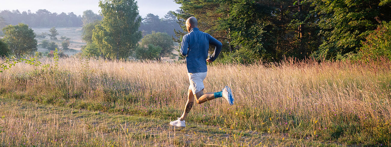 Man wearing the Sports Achilles Support while on a morning run through a grassy field. Man wearing Bauerfeind's Sports Achilles Support while on a morning run through a grassy field.
