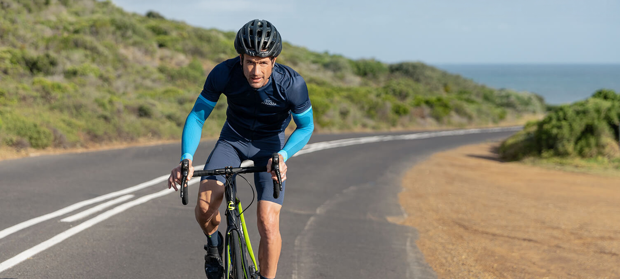 Cyclist wearing Bauerfeind's Sports Compression Arm Sleeves while riding uphill on a paved road surrounded by the ocean and grassy fields.