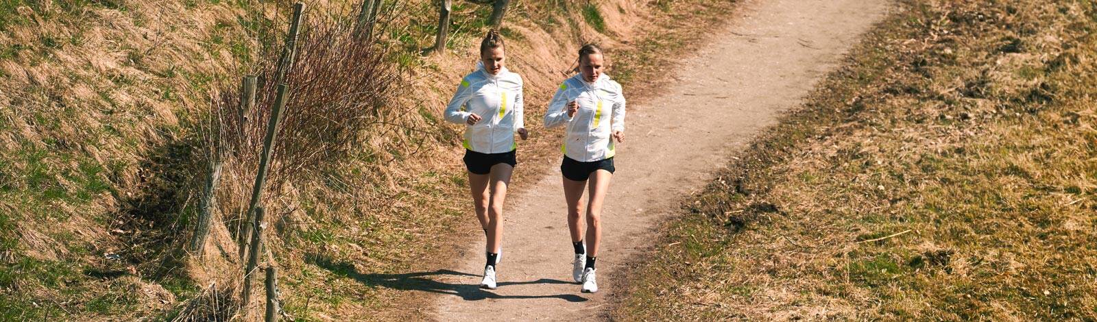Two women jogging on a dirt path. Banner image of two women jogging on a dirt path in a rural setting, dressed in white and yellow athletic wear.
