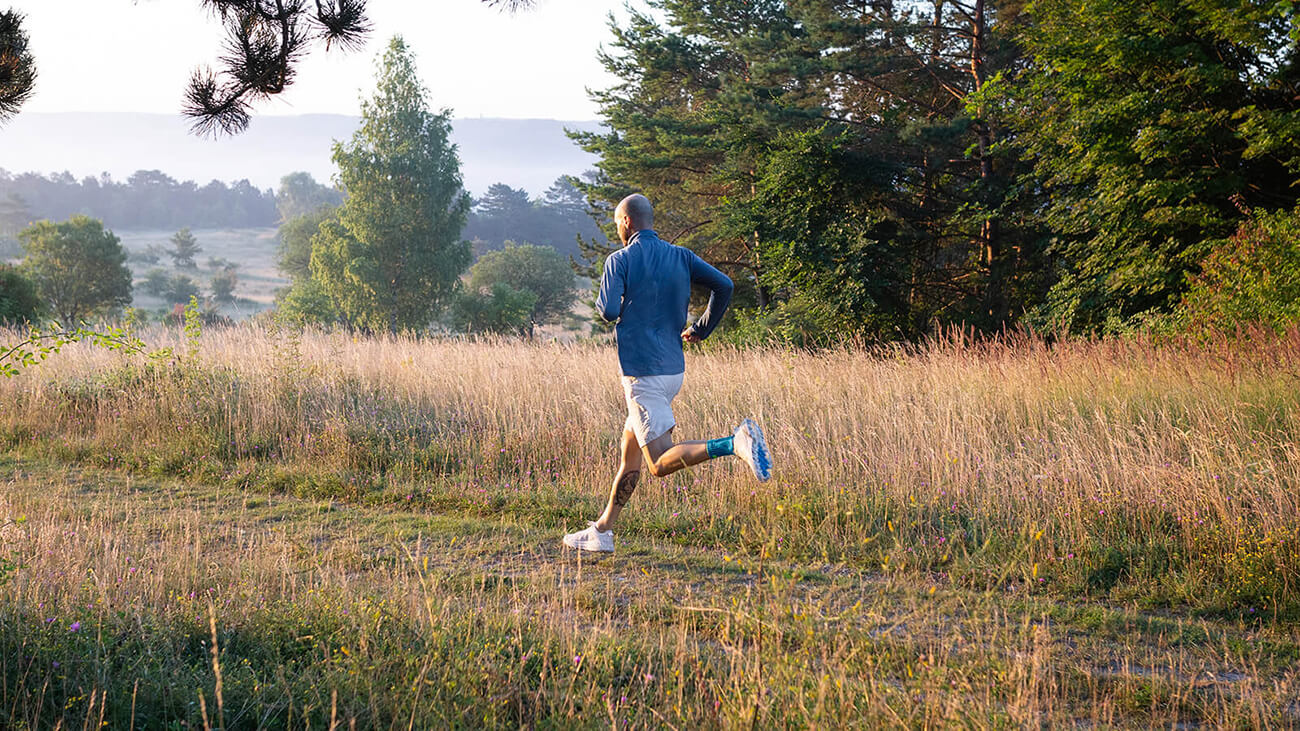 Wear the Sports Achilles Support for Enhanced Support and Relief While Running Man wearing Bauerfeind's Sports Achilles Support while on a morning run through a grassy field.