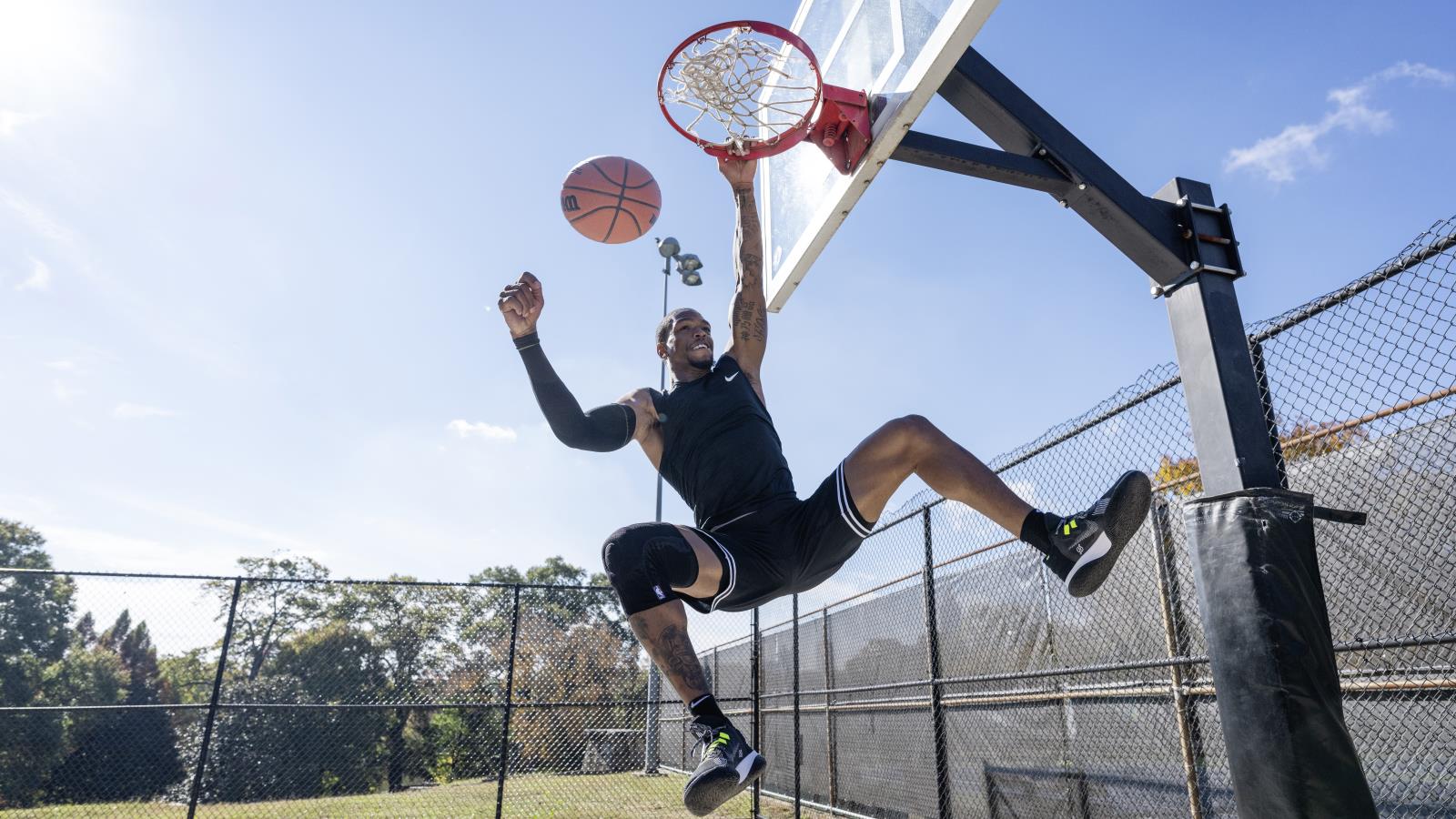 Athlete wearing the Sports Knee Support NBA while dunking on an outdoor basketball court. Athlete wearing Bauerfeind's Sports Knee Support NBA while dunking on an outdoor basketball court.