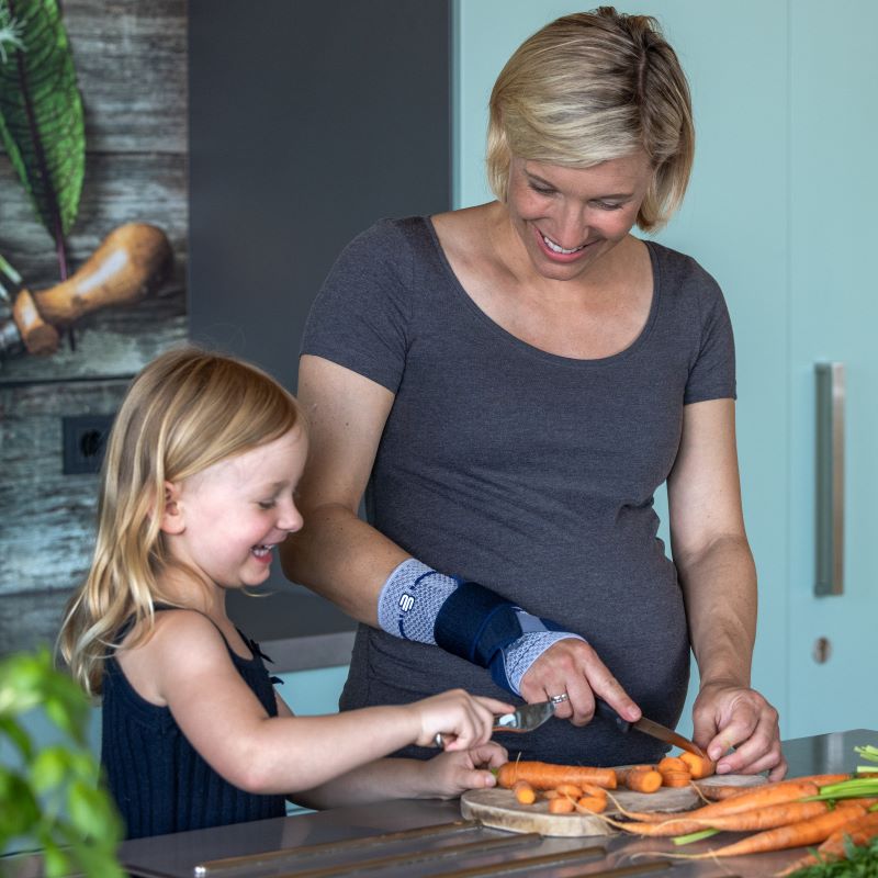 Comfortably Worn During Everyday Tasks Image of a woman wearing Bauerfeind's ManuTrain wrist brace (titan) while cutting carrots in the kitchen with her daughter.