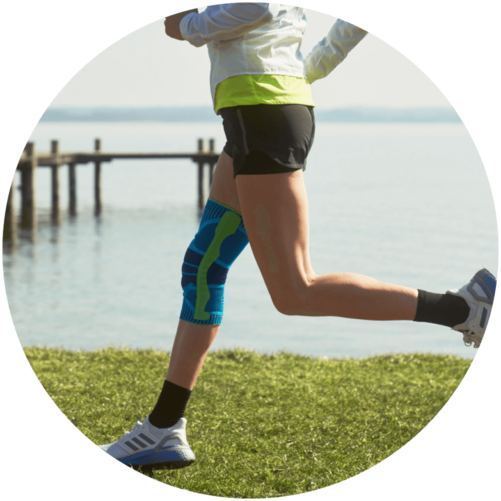 Durable Woman running on grass beside a lake while wearing Bauerfeind's Sports Knee Support.