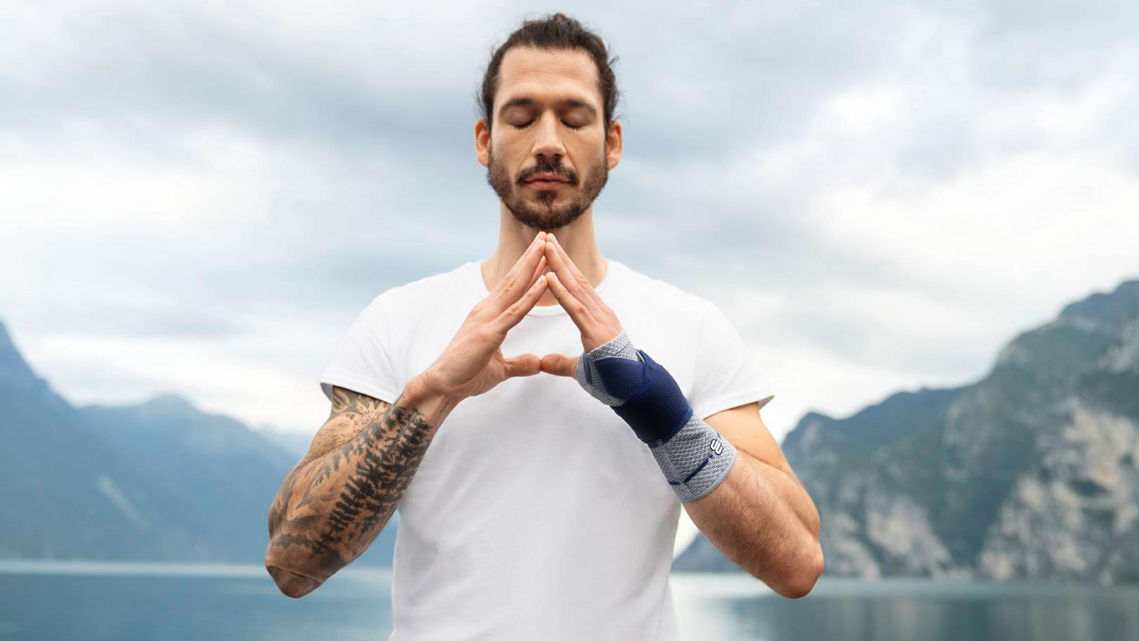 Man wearing the ManuTrain while practicing yoga and mindfulness meditation. Man wearing Bauerfeind's ManuTrain wrist brace while practicing yoga and mindfulness meditation outdoors near a lake and mountains.