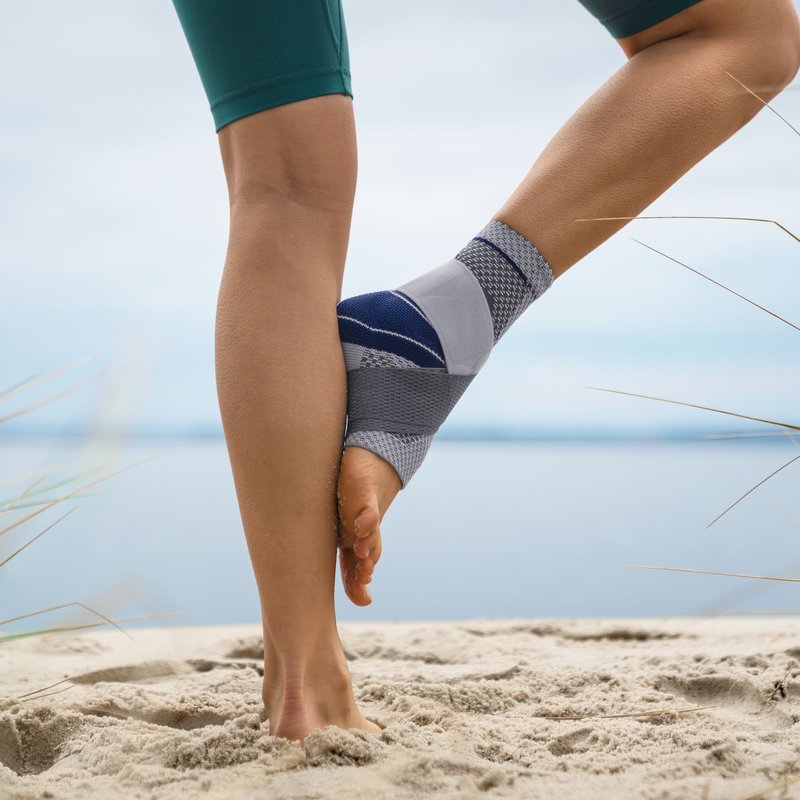 Close-up view of a person stretching on a sandy beach, wearing a grey and blue knee support brace and teal athletic shorts, with a calm lake and sky in the background.