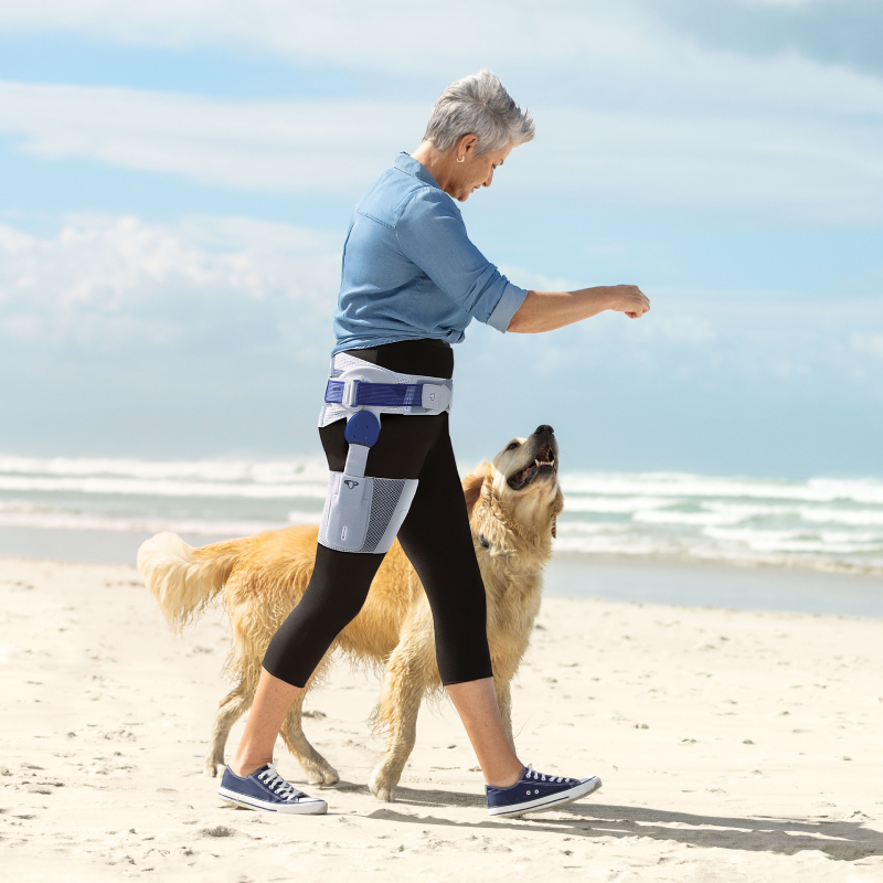 Woman wears Bauerfeind's CoxaTrain osteoarthritis hip brace while walking dog on the beach.