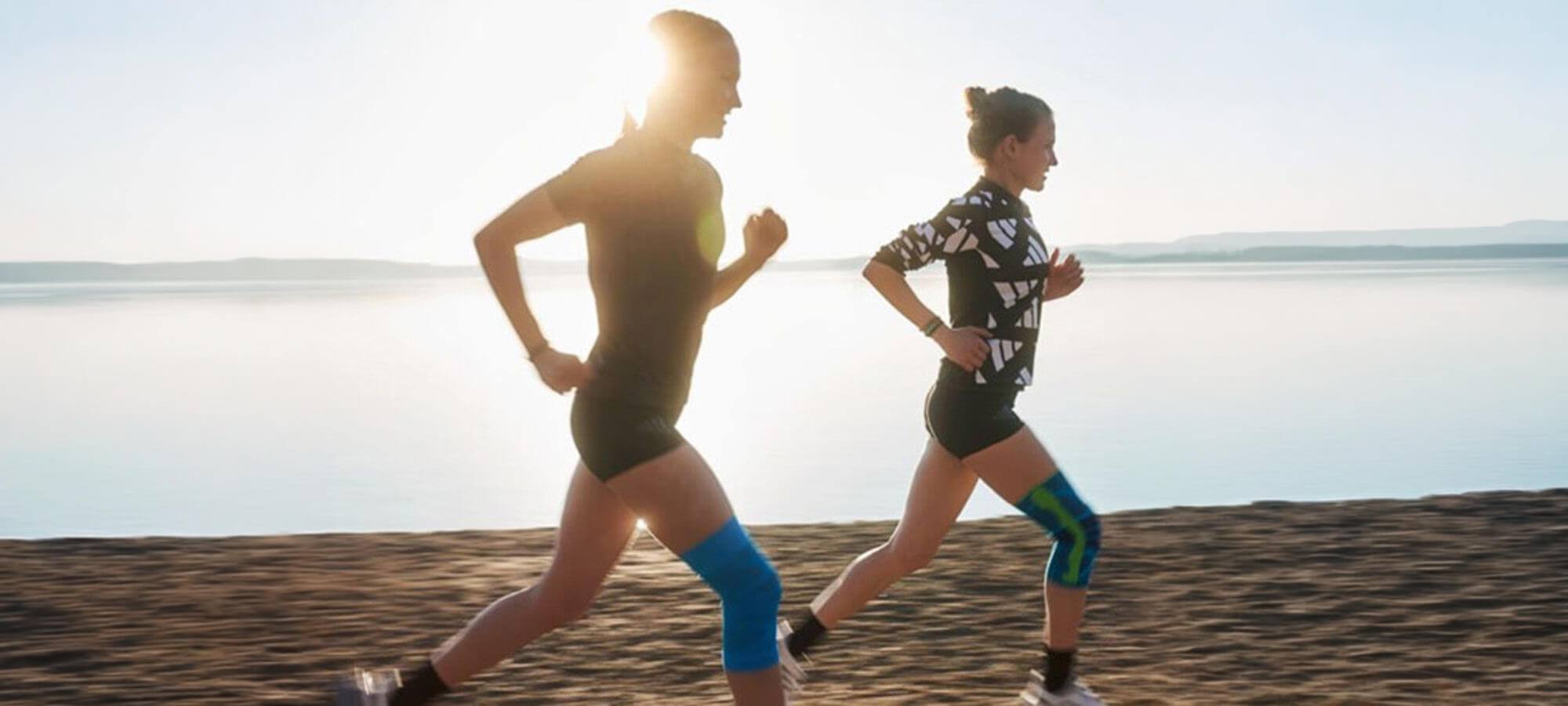 Two women running near a lake, with the runner on the left wearing Bauerfeind's Sports Compression Knee Support and the runner on the right wearing the Sports Knee Support.