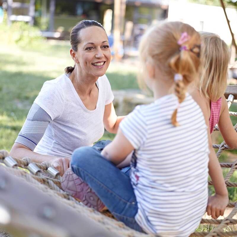 Woman wearing Bauerfeind's OmoTrain shoulder brace while playing at the park with two children.