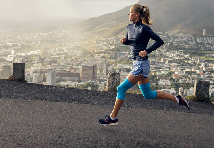 Run Further with Bauerfeind Woman wearing Bauerfeind's Sports Compression Knee Support while running on a road.