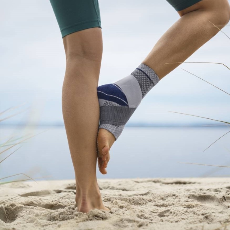 Exercise and Injury Prevention with Knee Support Brace at Beach Close-up view of a person stretching on a sandy beach, wearing a grey and blue knee support brace and teal athletic shorts, with a calm lake and sky in the background.