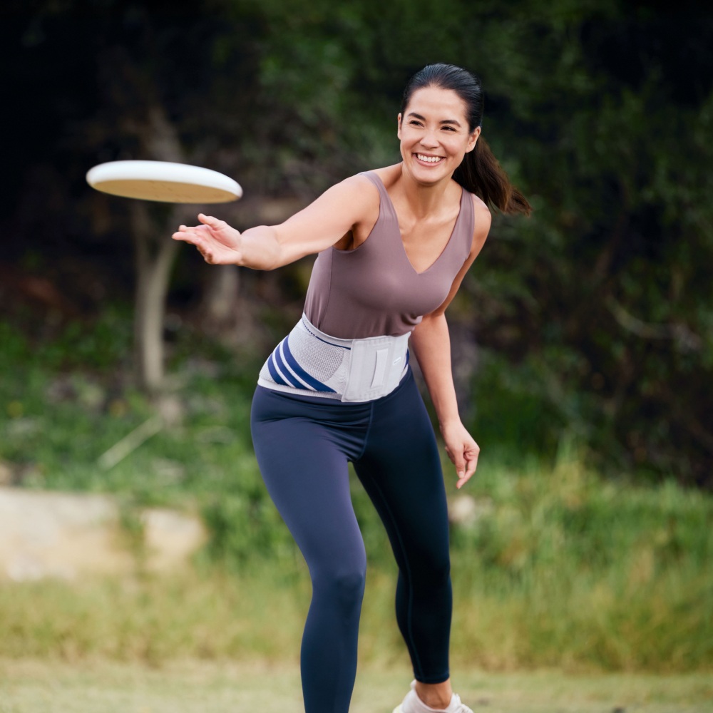 Woman wearing Bauerfeind's LumboTrain waisted back brace while throwing a frisbee in the park.