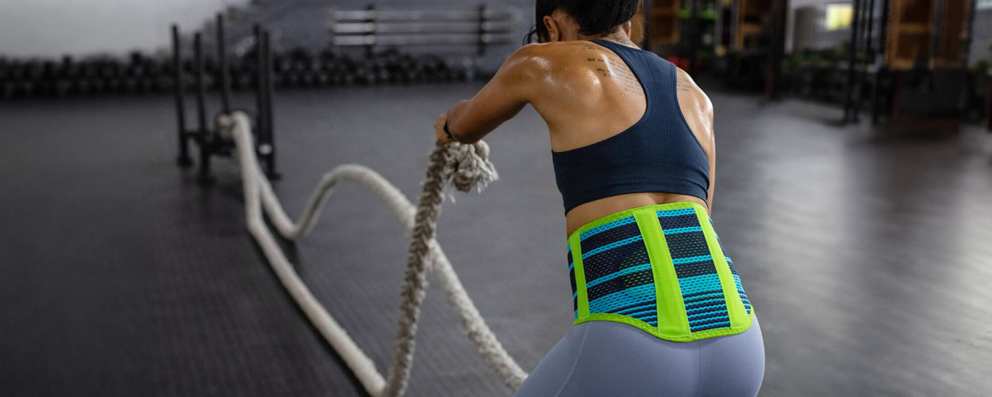 Woman wearing Bauerfeind's Sports Back Support while performing a rope exercise in an indoor gym.