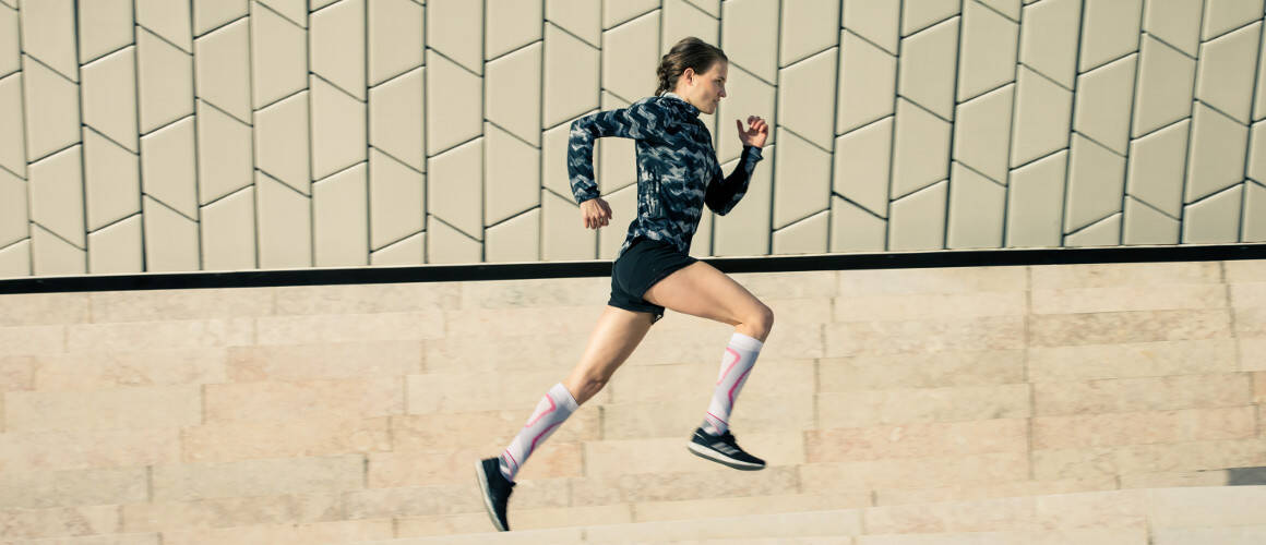 Woman wearing the Run Performance Compression Socks while running through a city street. Woman wearing Bauerfeind's Run Performance Compression Socks while running through a city street.