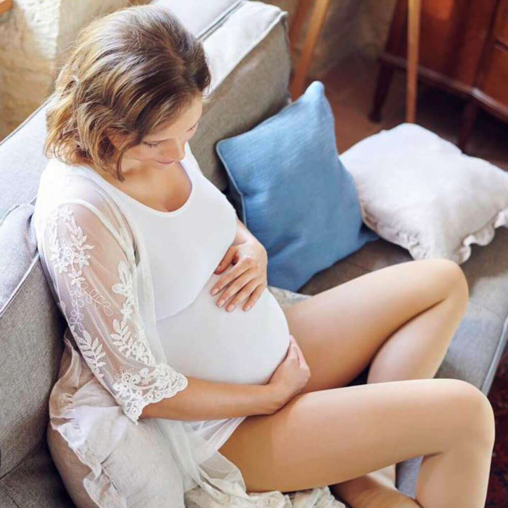 Pregnant woman sitting on a sofa while wearing the VenoTrain micro Compression Stockings. Pregnant woman in white dress sitting on a sofa and wrapping her arms around her belly while wearing Bauerfeind's VenoTrain micro Compression Stockings.