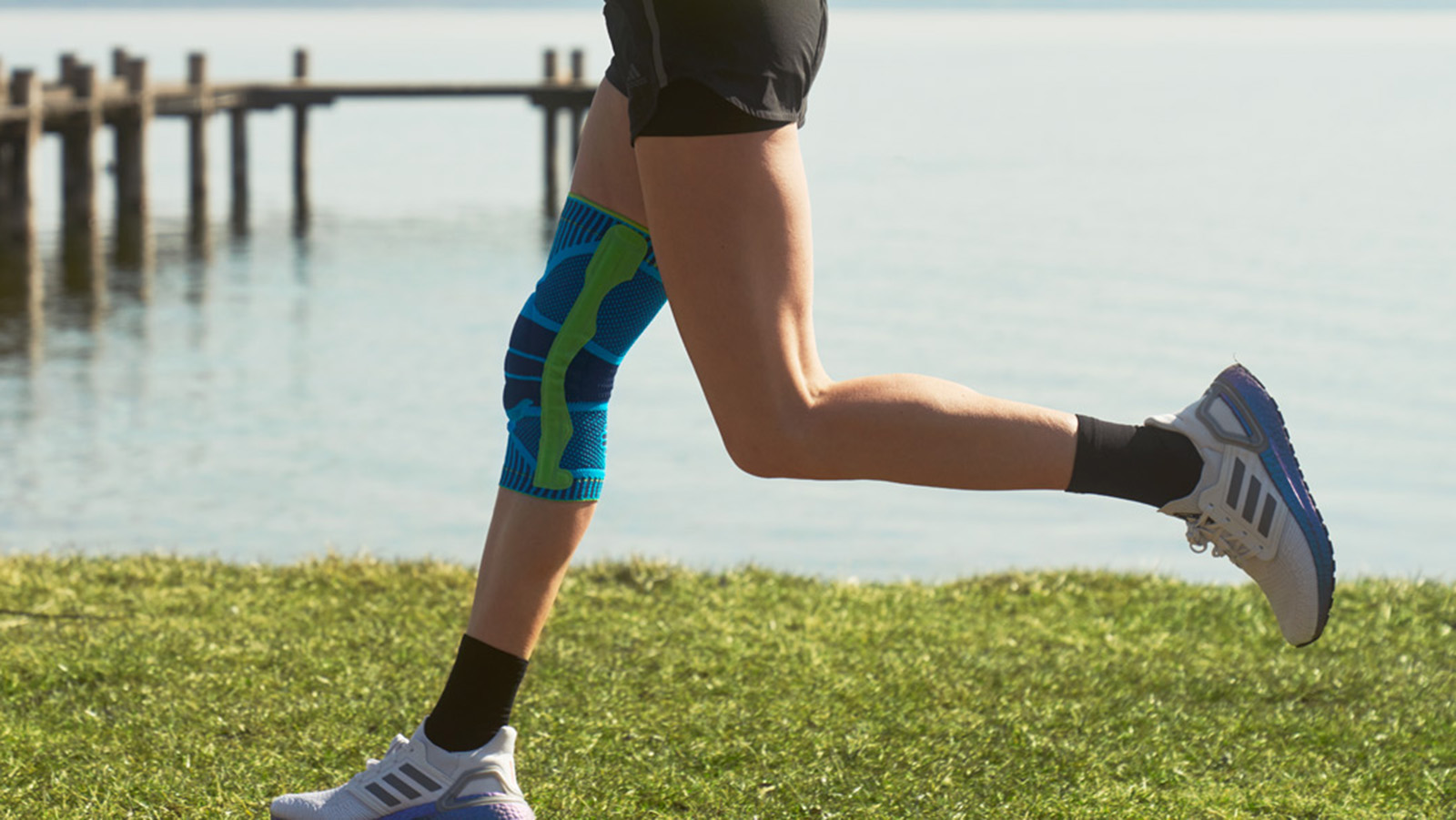 Bauerfeind's Sports Knee Support Woman wearing Bauerfeind's Sports Knee Support while running next to a lake.
