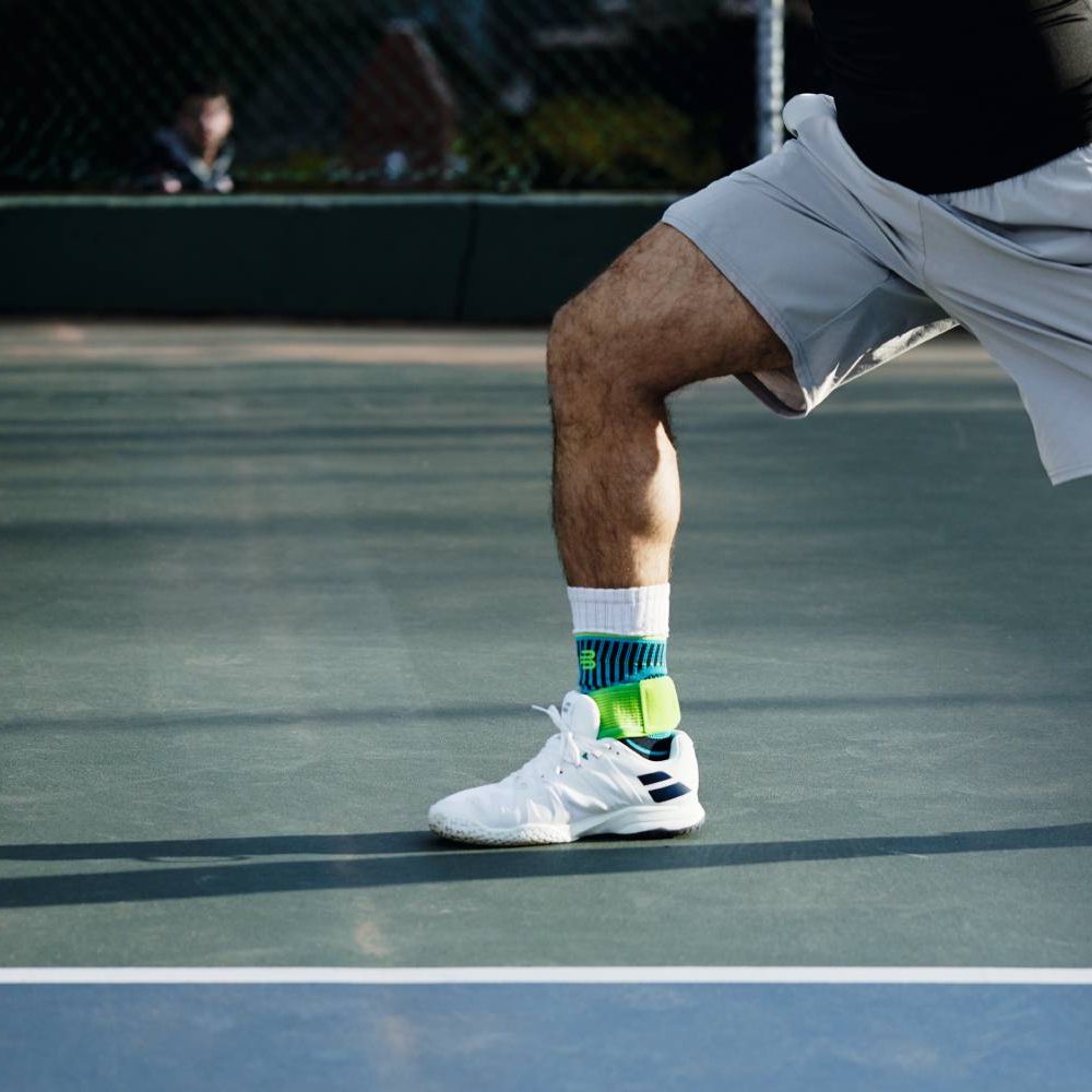 Close-up of a person wearing white sport shoes with neon green ankle supports, on tennis court
