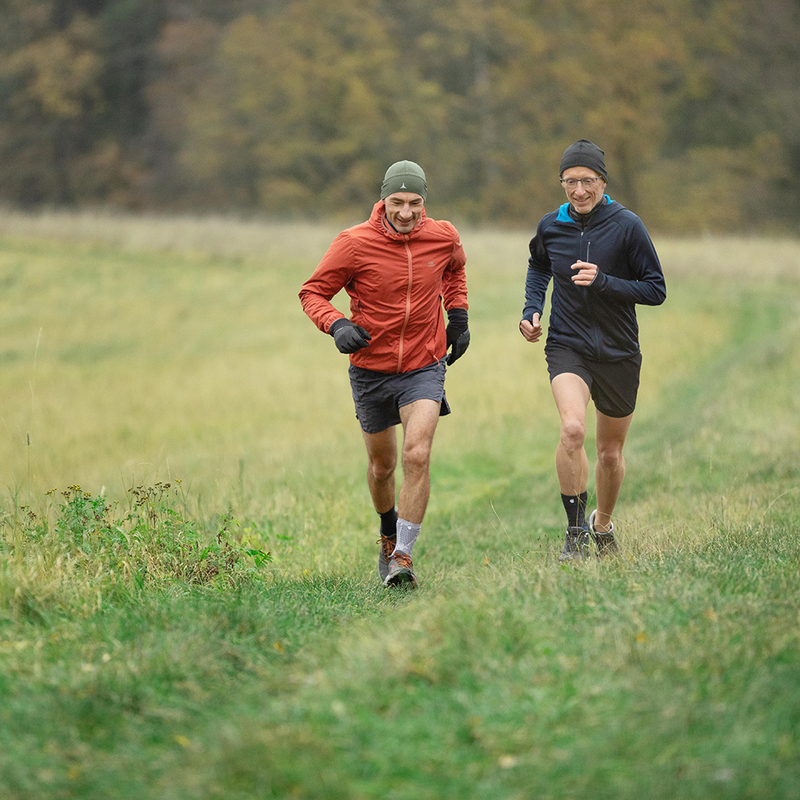 Outdoor Fall Jogging - Men Running in Athletic Wear on a Field Two adult men jogging in a field during fall, one wearing a red jacket and one in a blue jacket, both in exercise gear on a cloudy day.