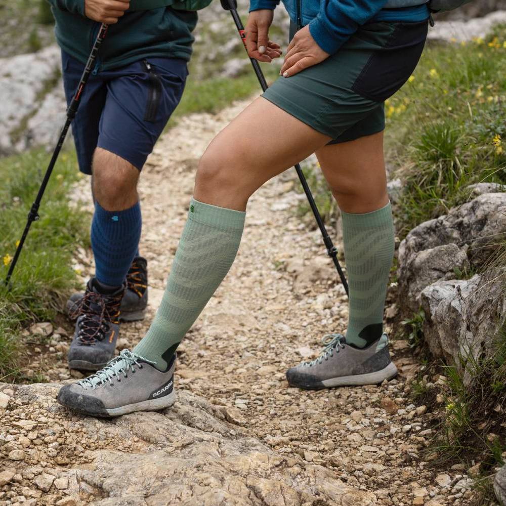 Two hikers wearing sturdy hiking boots and compression socks, using trekking poles on a rocky mountain trail.