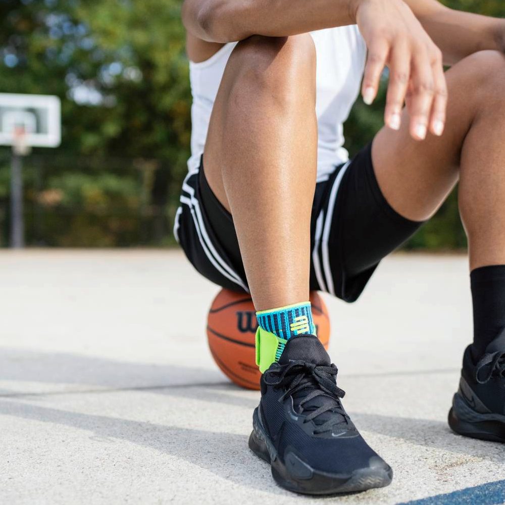 Basketball player wearing black sneakers and a blue ankle support sitting on an outdoor court with a ball