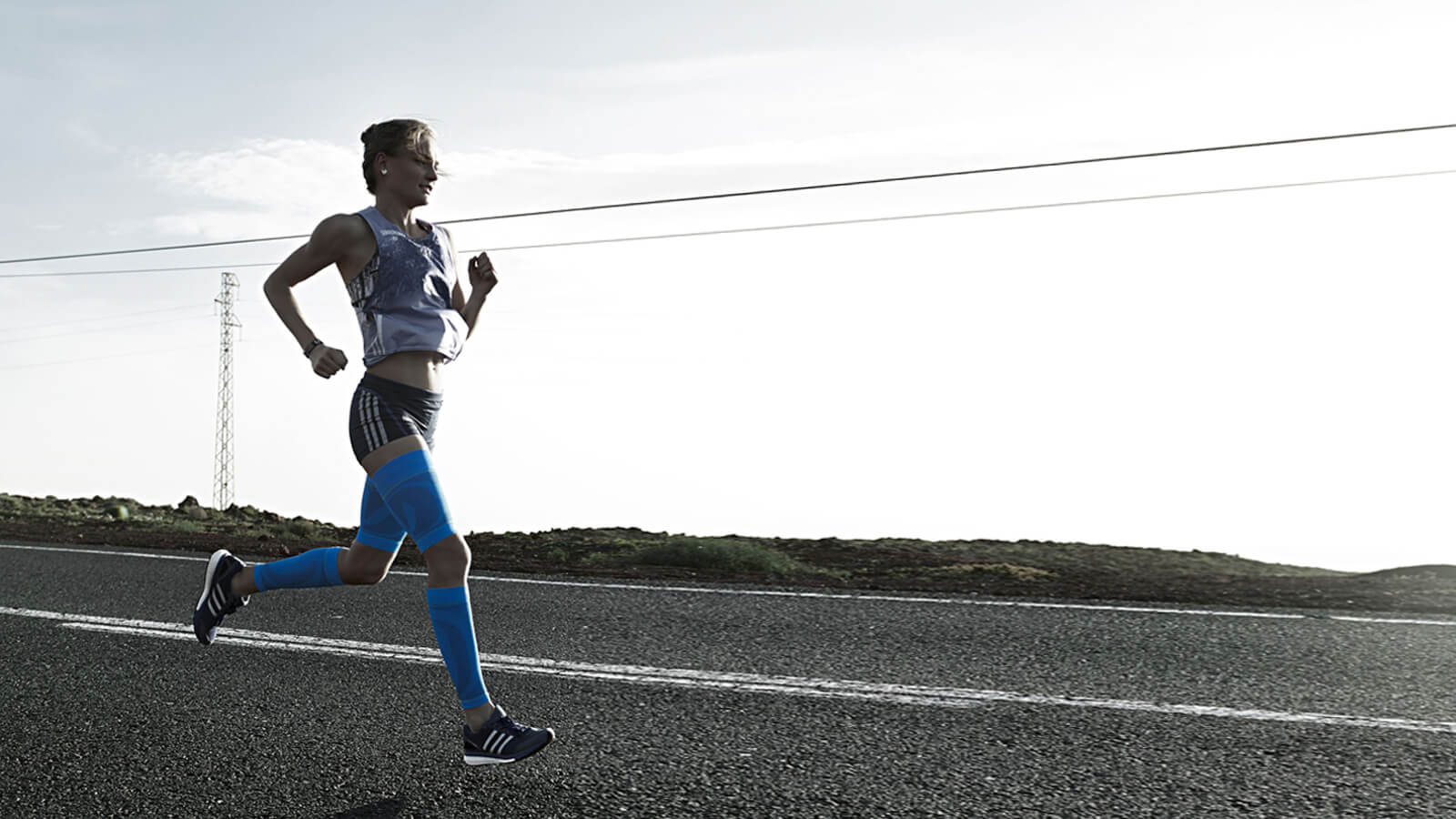 Woman running on a concrete road wearing Bauerfeind's Sports Compression Upper and Lower Leg Sleeves. Woman running along a concrete road while wearing Bauerfeind's Sports Compression Upper Leg Sleeves and Sports Compression Lower Leg Sleeves.
